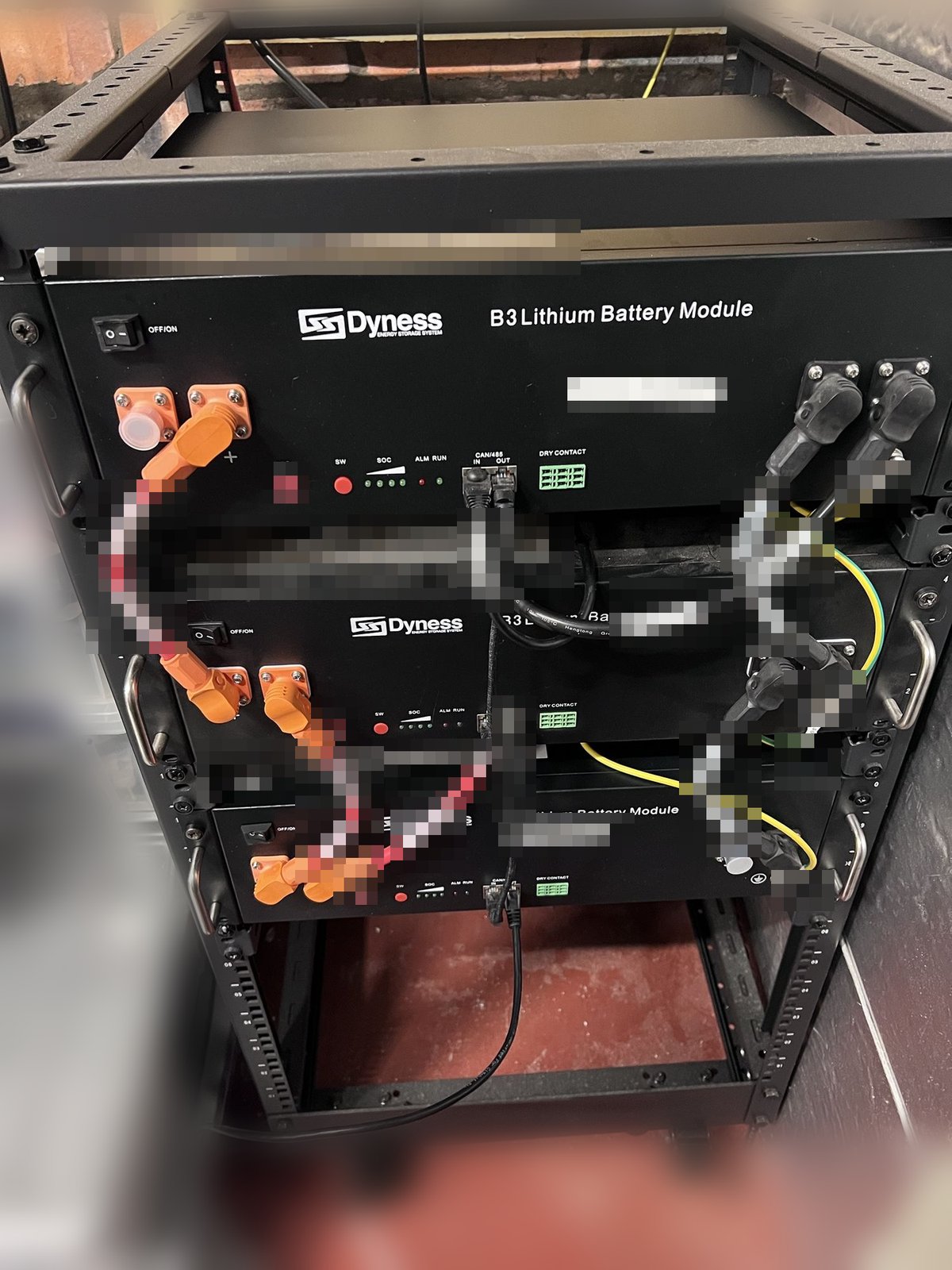 The rebuilt rack: a new Dyness B3 module at the top and bottom with the four-year-old one in the middle, orange DC links and the diagonal inverter feed, CAN patch leads chained IN to OUT, and the Pi's lead leaving the bottom module's OUT port. Serial stickers are blurred